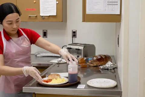 Student in a kitchen with apron and gloves, with a plate of bread, scrambled eggs and tomatoes