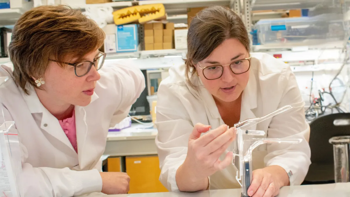 Katharina Maisel and Jenna Mueller in white lab coats looking at an applicator in the lab