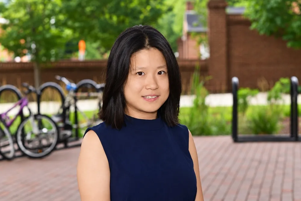 A photo of Suyue Cao standing outside. A bike rack, trees, and brick walls are in the background. 